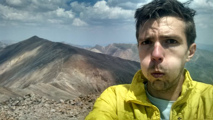 A tired hiker standing atop Sunshine Peak with Redcloud Peak in the background