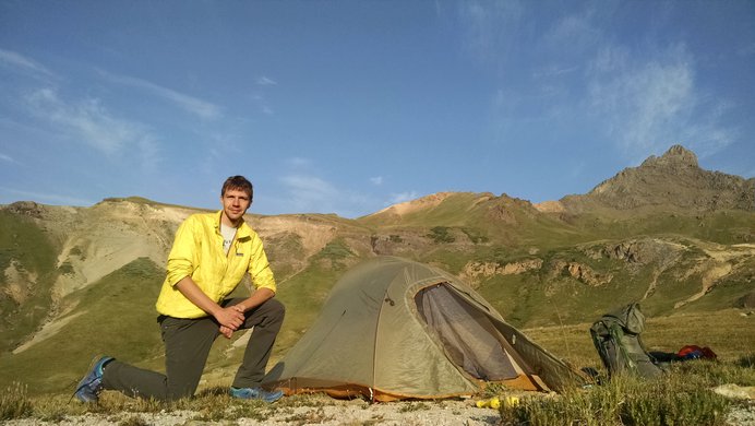 A hiker kneels in front of his tent in the Matterhorn Creek basin with Wetterhorn Peak in the background