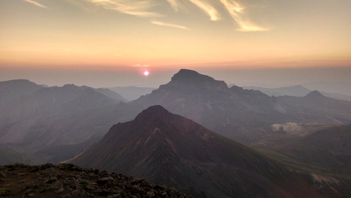 The sun begins to rise into a hazy sky behind Uncompahgre Peak, seen from Wetterhorn Peak